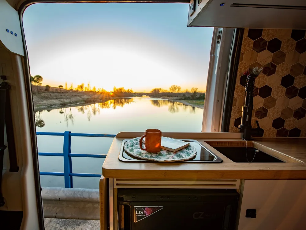 Cocina abierta de una camper Alquicamper con cafetera y vista a un lago al atardecer