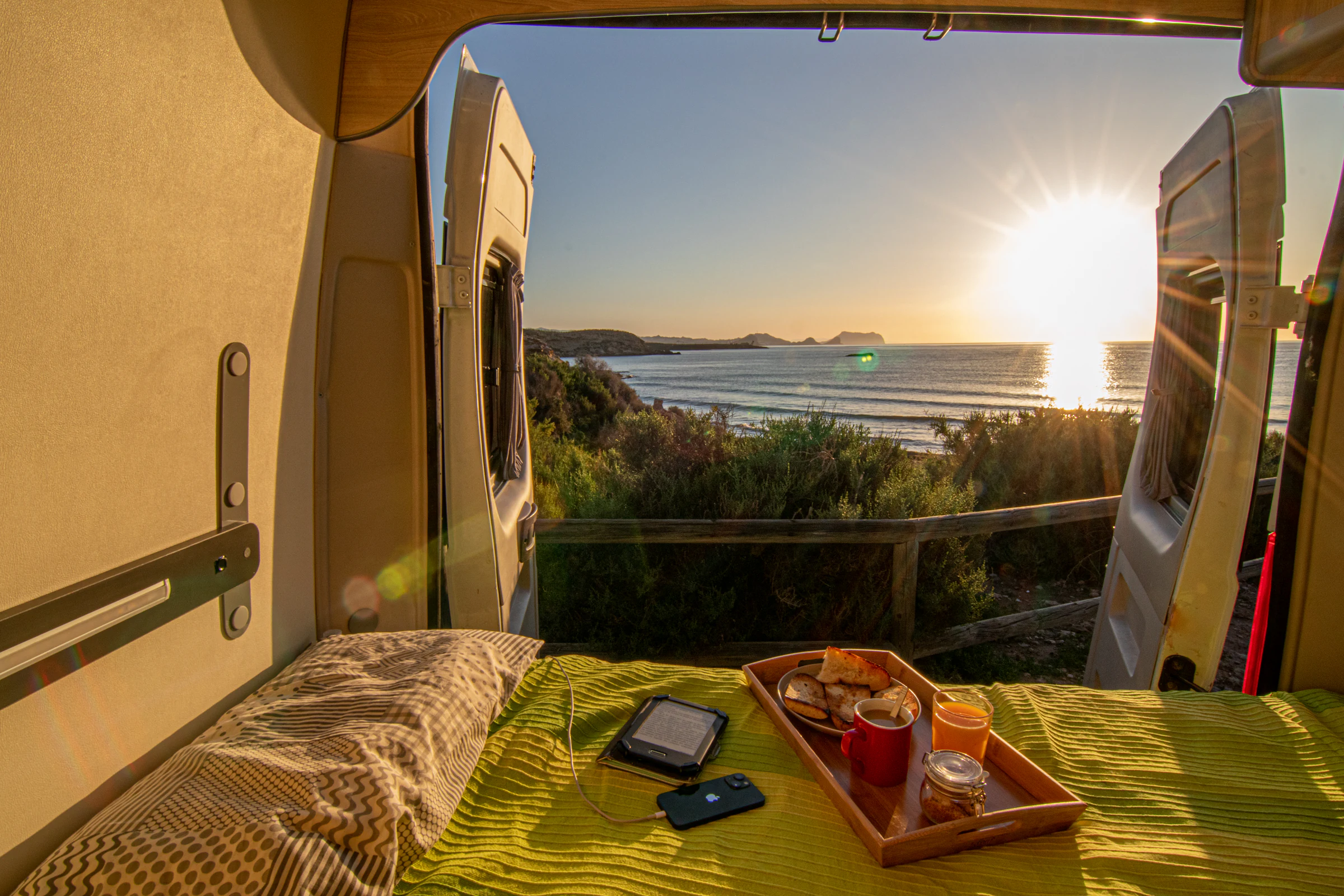 Vista al atardecer desde el interior de una camper Alquicamper · cama hecha y desayuno frente al mar · alquila tu camper en Madrid