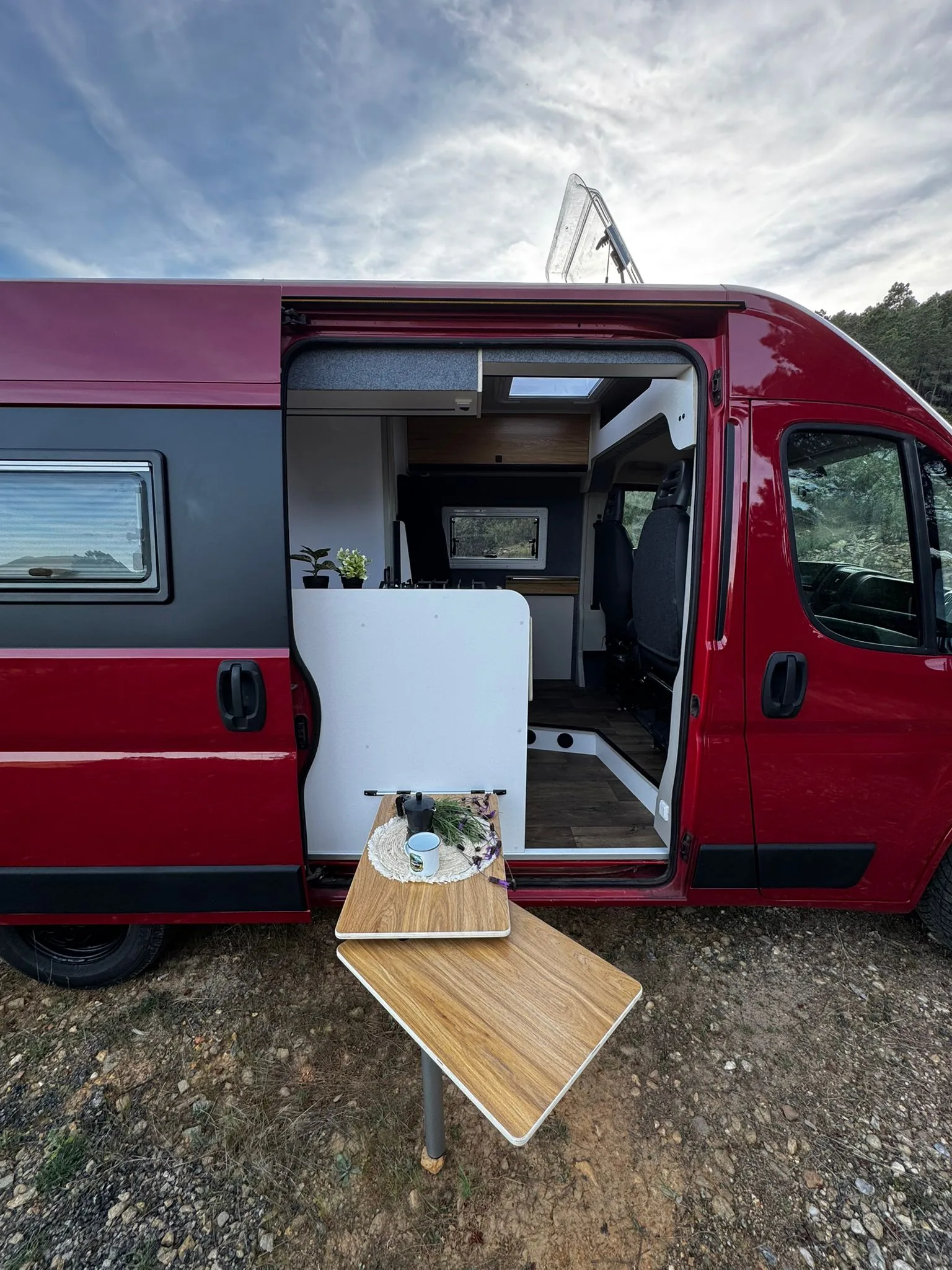 Camper Alquicamper roja con puerta lateral abierta y mesa exterior en un bosque