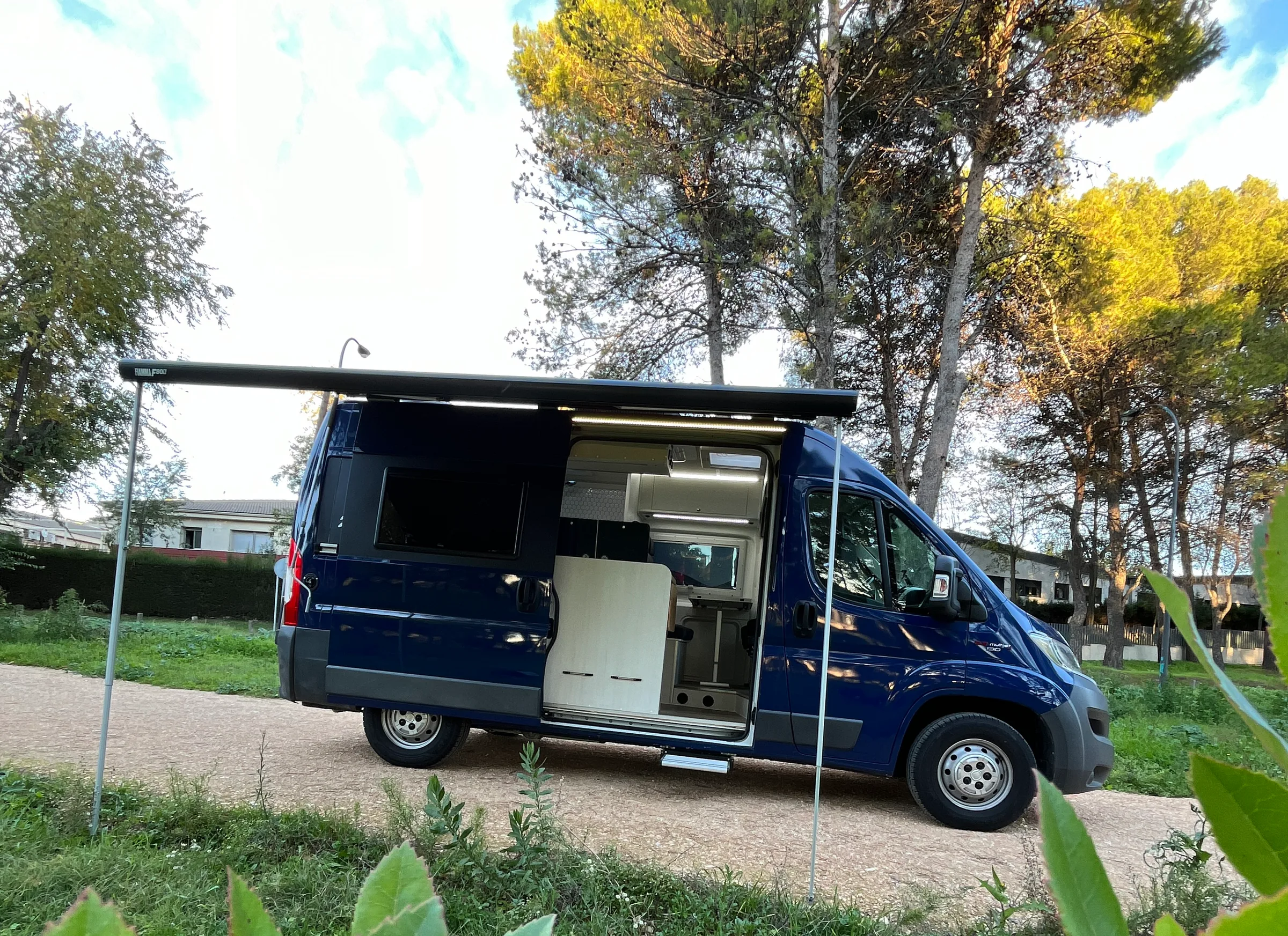 Camper Alquicamper azul con toldo extendido entre pinos · ruta camper en España