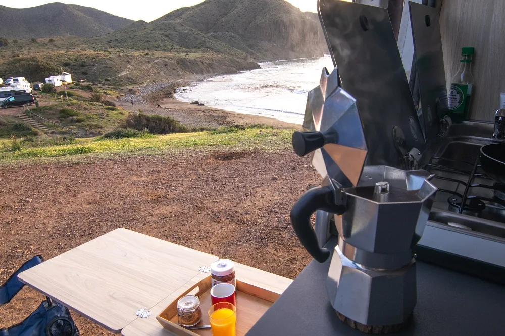 Desayuno con cafetera en la mesa exterior de una camper Alquicamper frente a una cala · verano en furgoneta camper