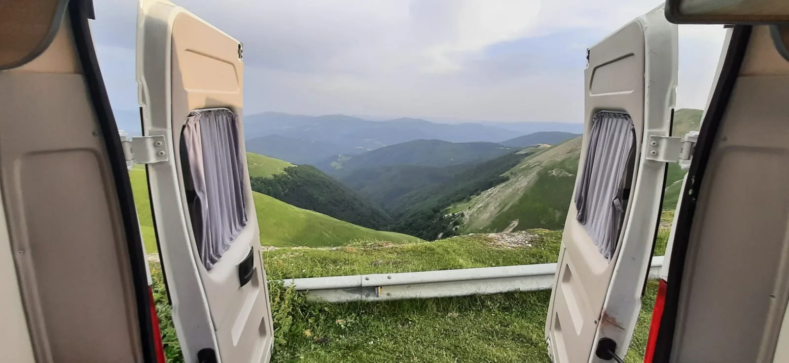 Puertas traseras de una camper Alquicamper abiertas a un valle verde de montaña · escapada de verano en camper