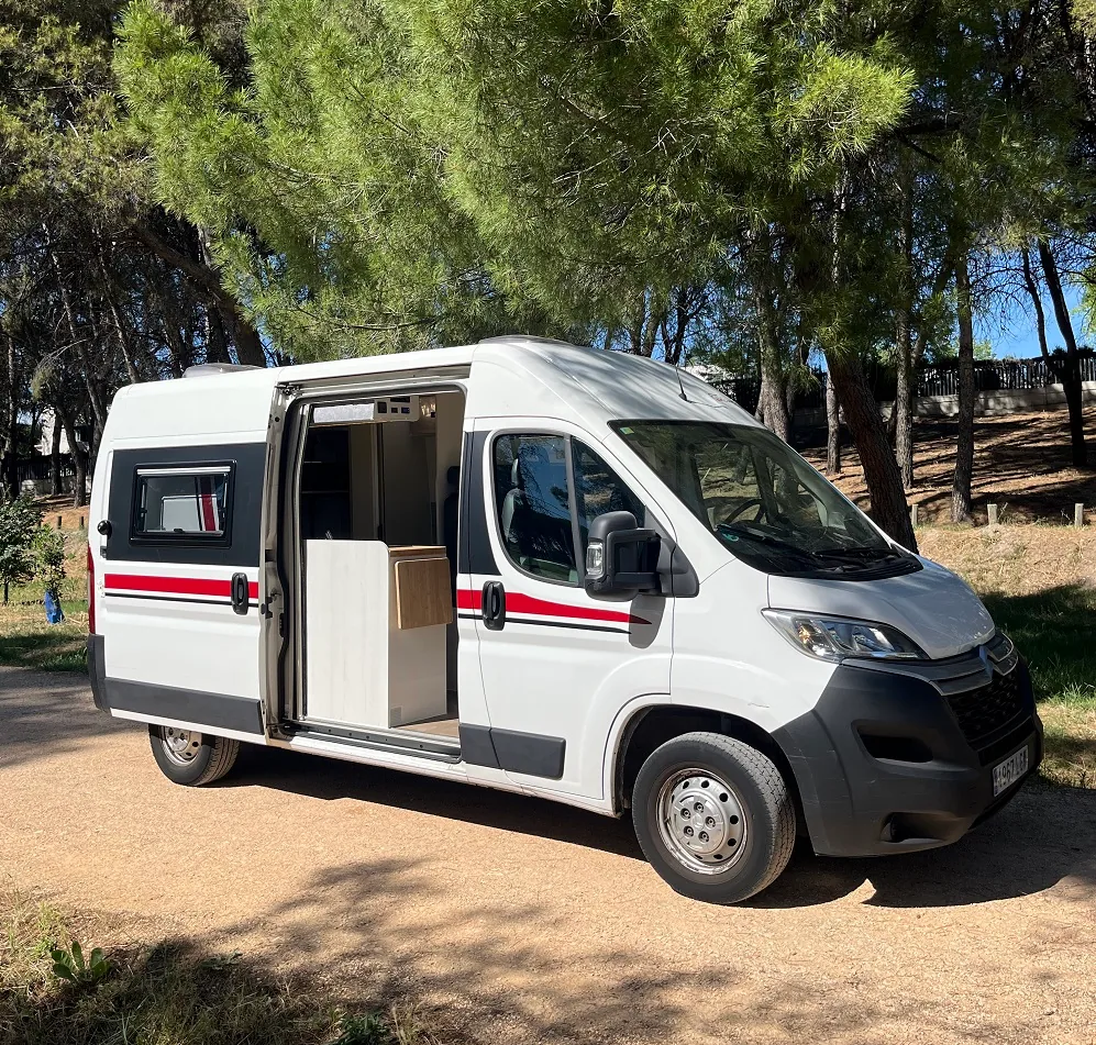 Furgoneta camper blanca con la puerta lateral abierta entre pinos, lista para alquilar en Madrid