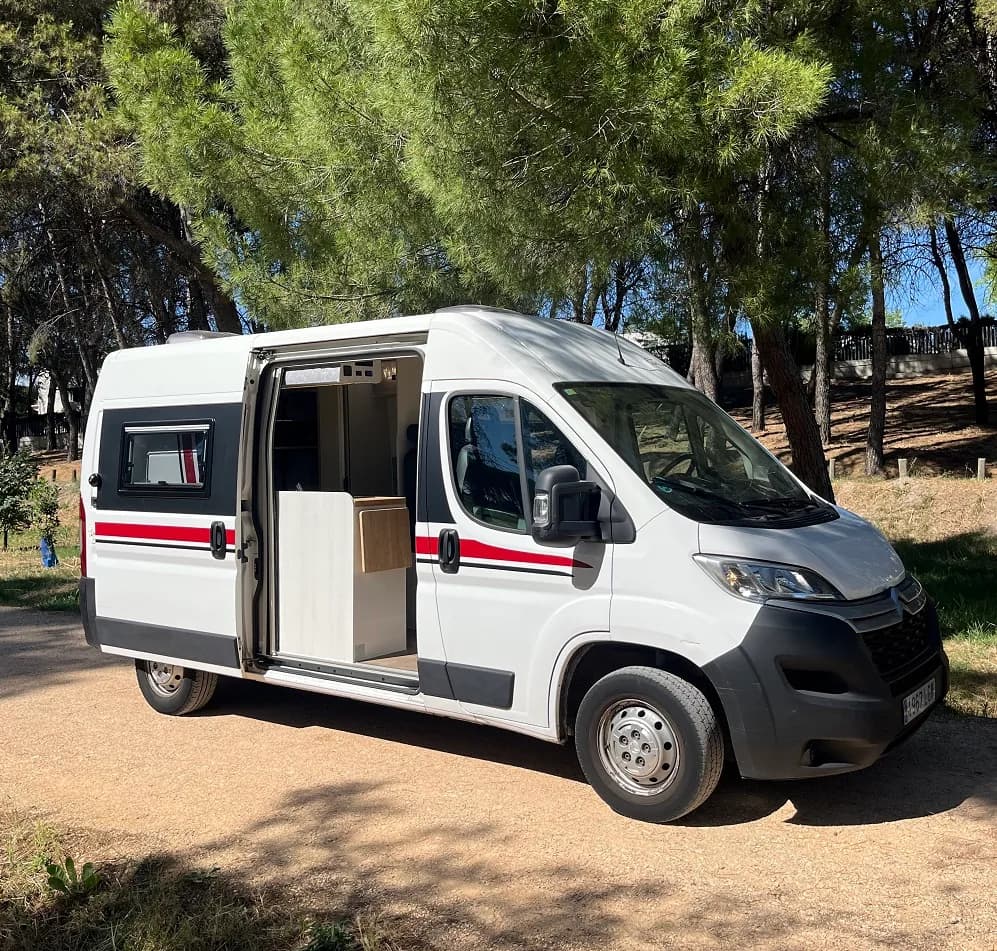 Furgoneta camper blanca con la puerta lateral abierta entre pinos, lista para alquilar en Madrid
