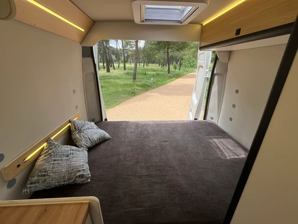 Interior de motorhome Alquicamper con cama hecha y vista al pinar · alquiler de casa rodante equipada para viajeros desde Latinoamérica